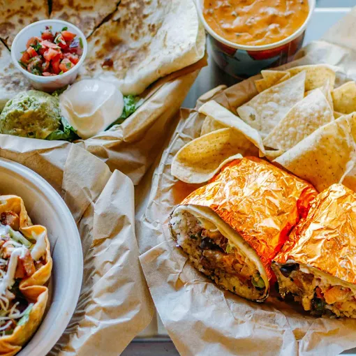 Fresh Mexican food spread featuring a foil-wrapped burrito, chips and queso, quesadilla, salsa, guacamole, and a taco bowl.