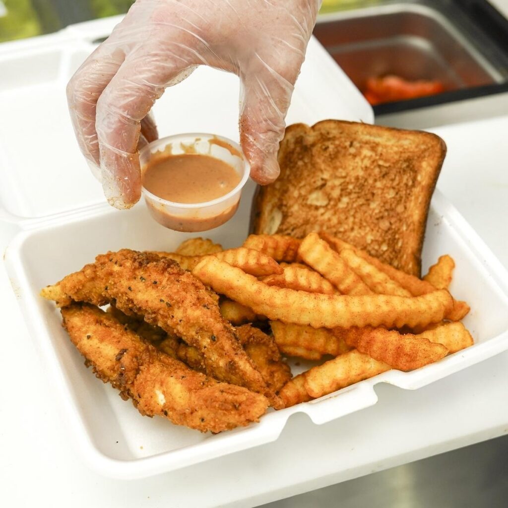 A takeout box filled with crispy seasoned chicken tenders, crinkle-cut fries, and Texas toast as a gloved hand holds a cup of dipping sauce.